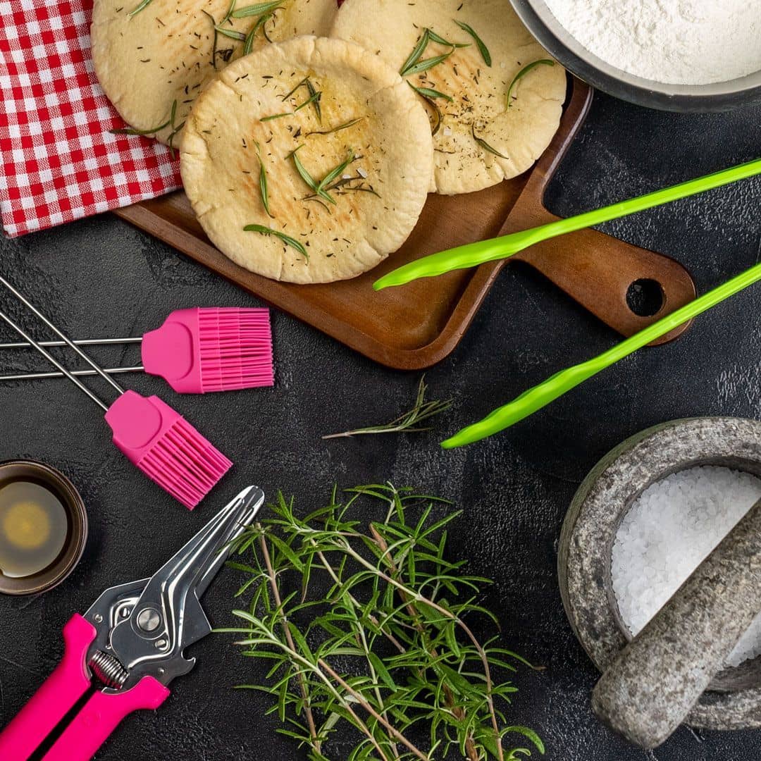 Flatbreads with rosemary on a wooden board, pink utensils, flour, oil, and salt on a dark surface.