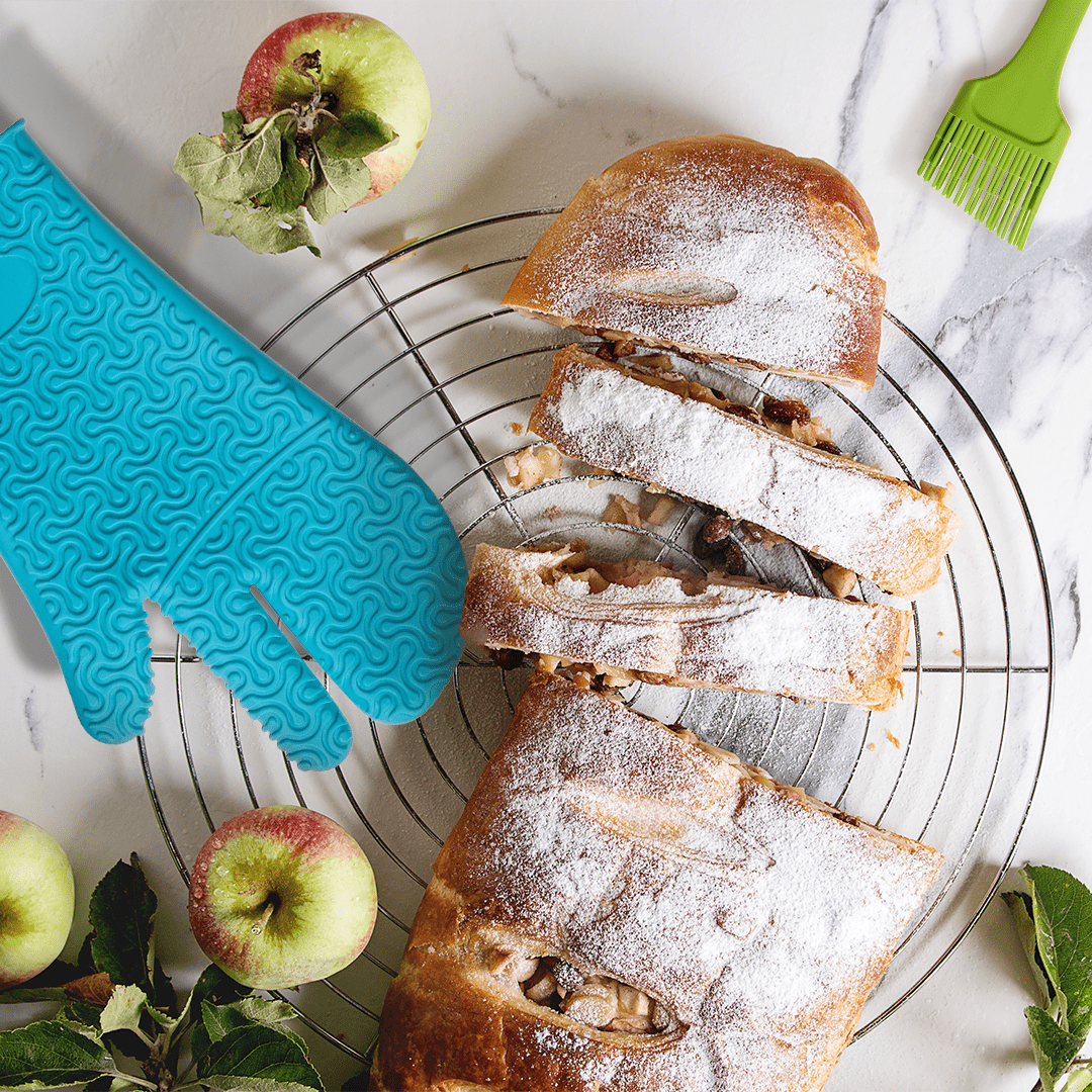Sliced apple strudel on a cooling rack with apples, a blue oven mitt, and a green pastry brush nearby.