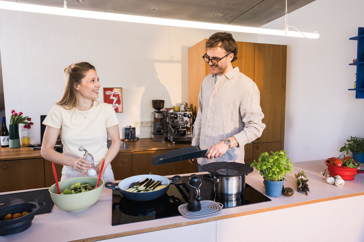Zwei Menschen, die lächelnd gemeinsam in einer modernen Küche Essen zubereiten, während auf der Arbeitsplatte frisches Gemüse liegt.