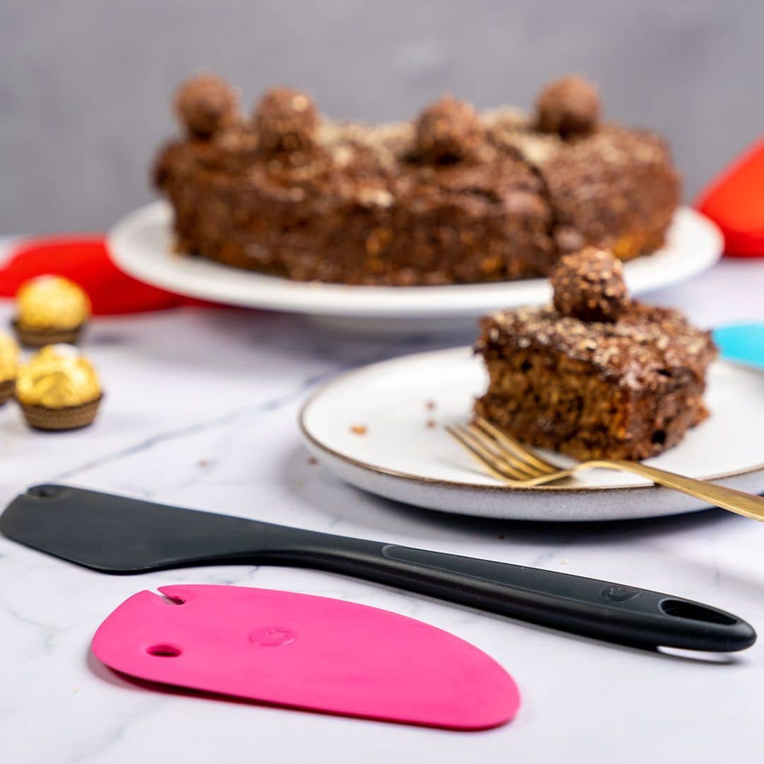 Chocolate cake with a slice on a plate, gold fork, spatulas, and chocolate candies on a marble surface.