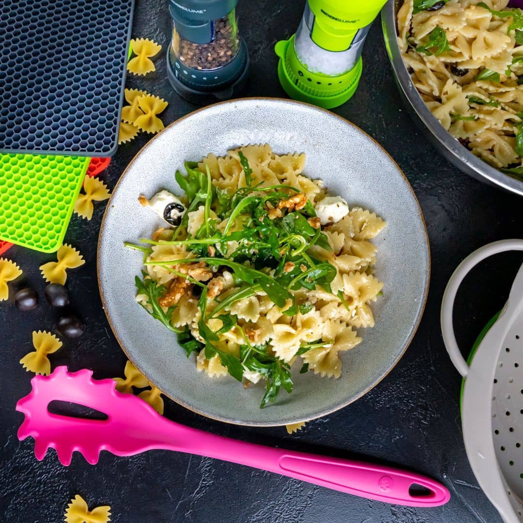 Bowl of farfalle pasta with arugula, olives, cheese, and walnuts, surrounded by kitchen tools and pasta.