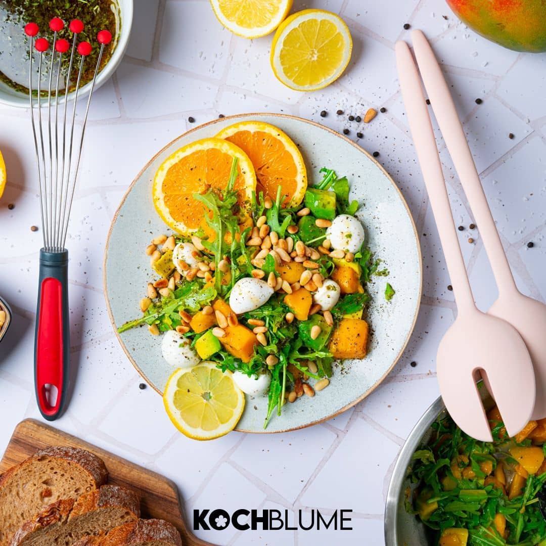 Fresh salad with greens, mozzarella, mango, pine nuts, and orange slices on a plate, with bread nearby.