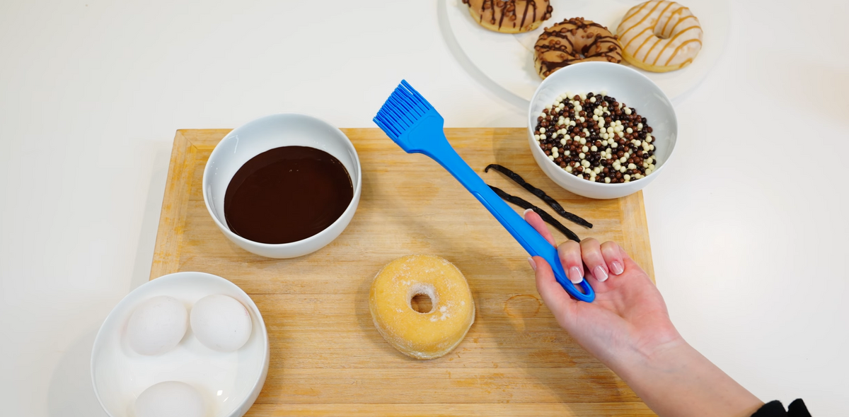 Eine Hand hält einen blauen Pinsel über einen Donut auf einem Brett, umgeben von Eiern, Schokolade und Streuseln.