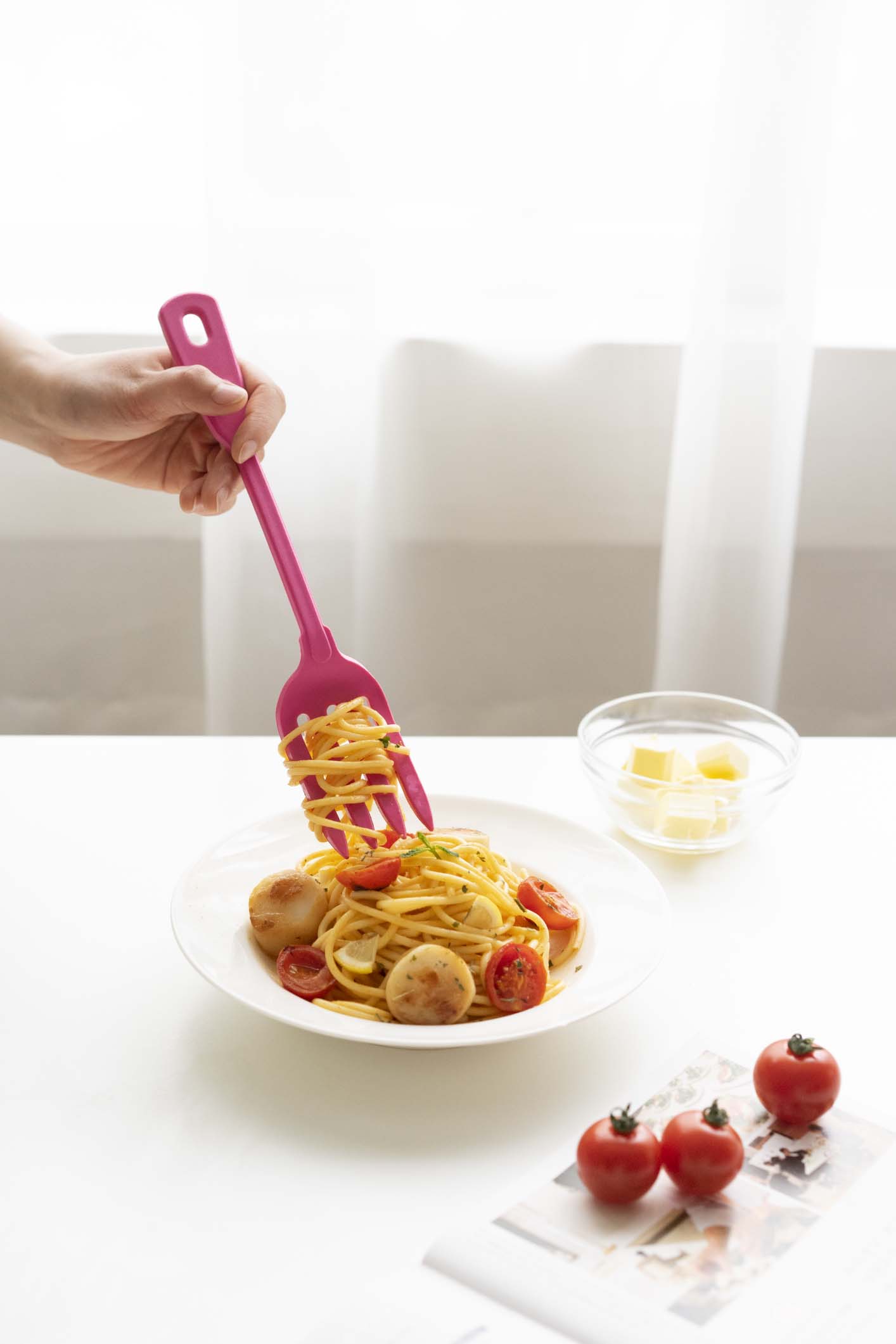 Hand hebt mit einer pinken Küchenzange Spaghetti und Würstchen auf einen Teller, neben Schüsseln mit Zutaten und frischen Tomaten auf einem hellen Tisch