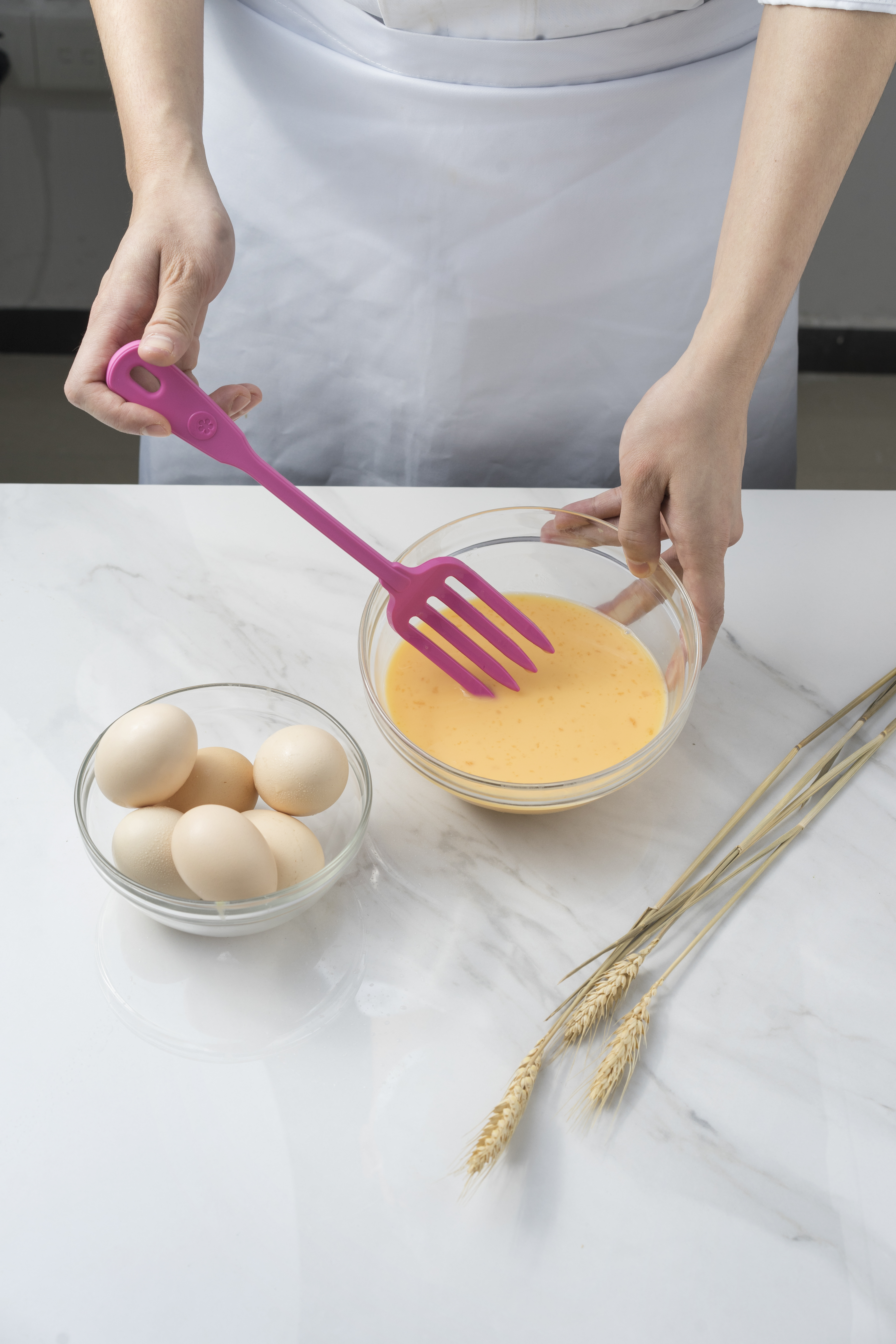 Eine Person schlägt Eiermischung in einer klaren Schüssel mit einem pinken Silikonschneebesen, daneben eine Schüssel mit ganzen Eiern und Weizenähren auf einer Marmoroberfläche