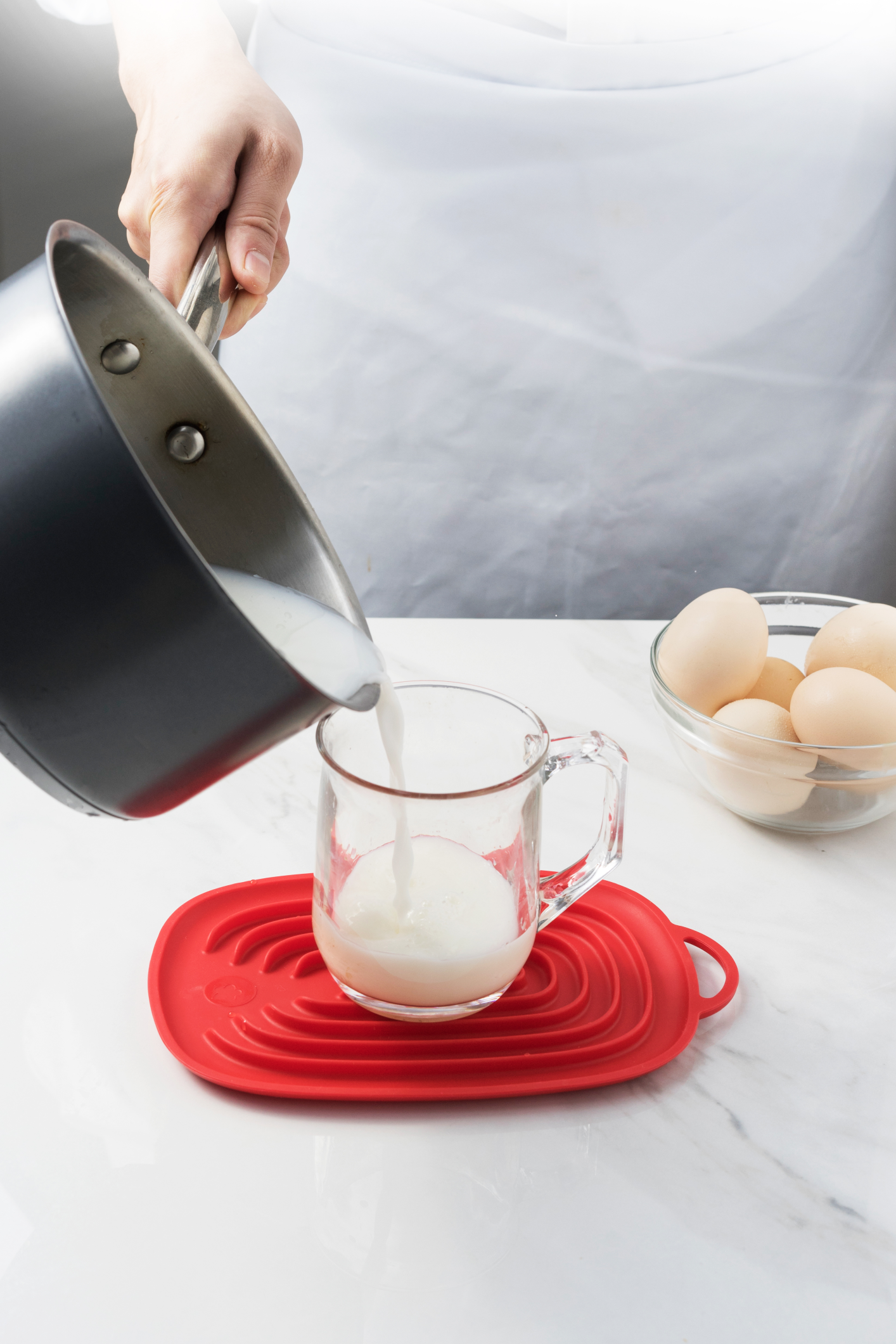 Eine Hand gießt Milch aus einem Topf in eine Messkanne auf einem roten Silikon-Untersetzer, auf einer Marmorarbeitsplatte mit Eiern im Hintergrund